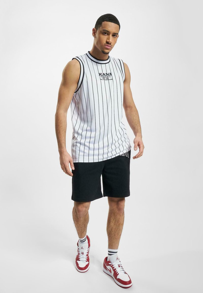 Hvid ærmeløs skjorte med sorte lodrette striber, sorte shorts og røde og hvide sneakers; minimal tekstdetalje på skjorten.