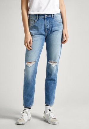 Light blue distressed jeans with rip details at knees, five-pocket design, and tapered leg. Worn with a white T-shirt and sneakers.