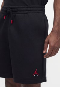 Shorts de sport noirs avec une taille élastique et un cordon, avec un petit logo Jumpman Air rouge et blanc sur la jambe gauche.
