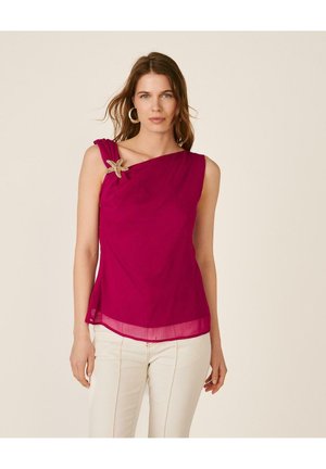 Mujer con top sin mangas de color magenta y broche en forma de estrella de mar en el hombro izquierdo, combinado con pantalones blancos sobre un fondo liso.