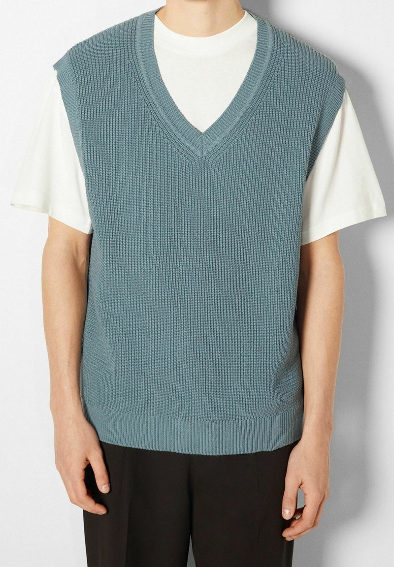 Jasnoniebieska dzianinowa kamizelka z dekoltem w serek, prążkowaną fakturą i krótkimi rękawami, noszona na białej koszulce, w połączeniu z czarnymi spodniami.