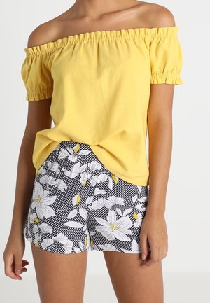 Vrouw draagt een gele off-shoulder top en zwart-witte shorts met bloemmotief en gele accenten tegen een effen achtergrond.