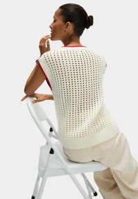 Maglione lavorato a maglia leggero di colore crema con motivo a cremiera e rifiniture rosse. Modello seduto su una sedia pieghevole bianca, che indossa pantaloni beige.