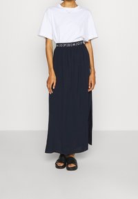 Jupe maxi bleue marine avec une taille froncée ornée d'une bande logo noir et blanc. Associée à un t-shirt blanc ample et des sandales noires.