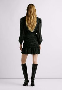 Mini vestido negro con textura, mangas abullonadas, cintura ajustada y dobladillo con volantes. Complementado con botas de goma negras de caña alta y brillante.