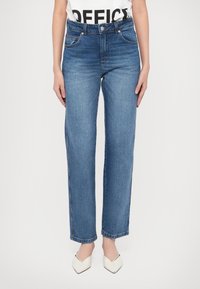 Femme portant un jean bleu à jambes droites, des chaussures blanches à bouts pointus et une chemise blanche partiellement visible avec un texte noir en gras.