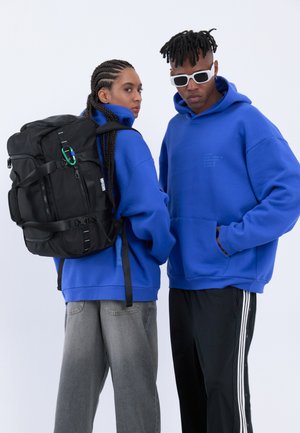 Due giovani adulti che indossano felpe con cappuccio blu, uno con jeans grigi e uno zaino nero, l'altro con pantaloni sportivi neri con righe bianche e occhiali da sole bianchi.