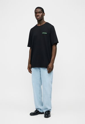 Homme portant un t-shirt noir oversize avec un petit texte vert, un jean bleu clair à jambes larges et des chaussures noires, debout devant un fond blanc.