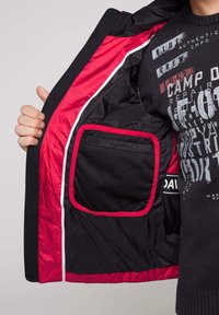 Chaqueta negra con un forro de satén rojo, que presenta un bolsillo visible con textura de malla. El interior tiene un ribete rojo que contrasta.
