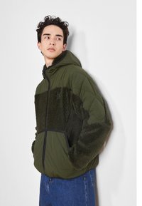 Veste matelassée vert olive avec un devant en polaire texturée. Elle possède une capuche, une fermeture éclair et des poches latérales. Le design comprend des panneaux contrastés.