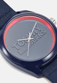 Reloj de pulsera azul marino con una correa de tela texturizada, que presenta una esfera gris grabada con "TOMMY JEANS" y un acento rojo alrededor del cristal.