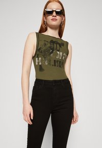 Λαδί ριγέ αμάνικο τοπ με γραφικό σχέδιο, συνδυασμένο με ψηλόμεσα μαύρα τζιν. Τα αξεσουάρ περιλαμβάνουν ορθογώνια γυαλιά και κρίκους σκουλαρίκια.