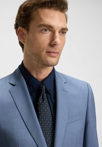 Jasnoniebieska marynarka o delikatnej fakturze, zestawiona z ciemną koszulą i krawatem w kropki. Posiada jedną kieszeń na piersi i klapy typu notch.