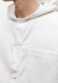 Camisa blanca con capucha que presenta una tapeta de botones, tela texturizada y un bolsillo en el pecho. El cuello es suave y tiene un diseño de corte relajado.
