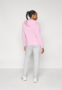 Ružičasta hoodie s kapuljačom i kengur džepom, u kombinaciji s svijetlosivim trenerkama. Oboje izrađeno od mekanog materijala, prikladno za svakodnevno nošenje.