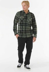 Camicia di flanella a quadri verde e nero con due tasche sul petto, abbinata a pantaloni neri e sneakers nere con dettagli bianchi.