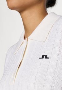 Polo shirt bianco lavorato a maglia con colletto a coste e logo nero ricamato sul petto. Presenta un motivo a treccia testurizzato.