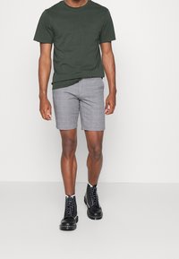 T-shirt en coton vert associé à un short à carreaux gris. Le mannequin porte des bottes noires à lacets avec une semelle blanche contrastante. Design minimaliste, sans motifs.