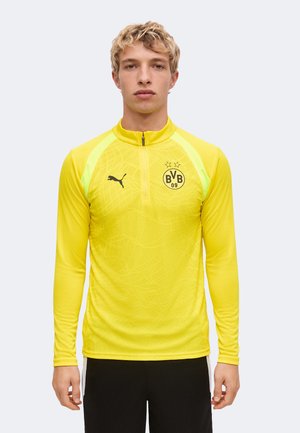 Blondyn stojący na jasnym jednolitym tle, ubrany w żółtą sportową koszulkę z długim rękawem z logo BVB 09 i Puma.
