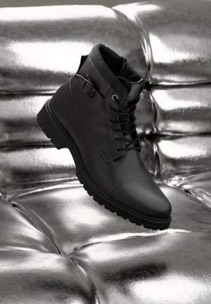 Bottes noires en cuir jusqu'à la cheville avec lacets, bout rond et semelle en caoutchouc. Présente une sangle avec boucle sur le côté et des accents texturés noirs.