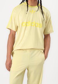 Camiseta Adidas de color amarillo claro con rayas texturizadas, logotipo amarillo en negrita y tres acentos de rayas en los hombros, combinada con pantalones a juego.