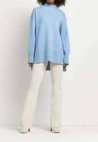 Suéter oversized azul claro con cuello alto, con detalles de botones en un hombro, combinado con pantalones de campana color crema y tacones claros.