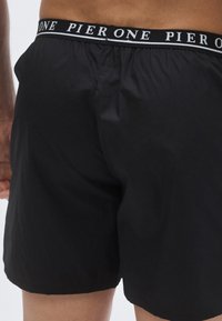 Shorts de bain noirs en tissu léger avec une ceinture élastique arborant un logo texte blanc "PIER ONE". Design basique, sans motifs.