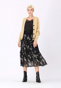 Cardigan en tricot beige avec des boutons noirs, superposé sur un débardeur en dentelle noire et une jupe noire à volants avec des motifs de plumes blanches. Bottines noires.