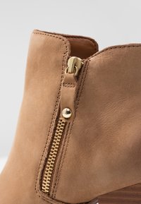 Stivale alla caviglia in pelle marrone con dettaglio della cerniera dorata e tacco in legno. Texture liscia con cuciture visibili lungo i bordi.