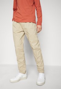 Chino beige con una vestibilità tapered e tasche laterali. Abbinato a una camicia a maniche lunghe di colore ruggine e sneaker bianche. Tessuto morbido.
