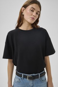 Jauna sieviete valkā vienkāršu melnu oversize T-kreklu, ieliktu zilās džinsās ar melnu ādas jostu un pērļu auskariem.