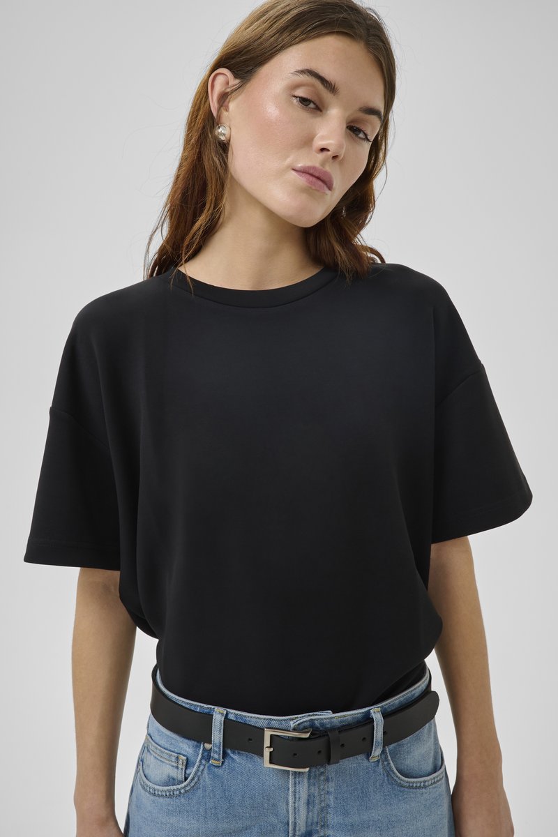 Jauna sieviete valkā vienkāršu melnu oversize T-kreklu, ieliktu zilās džinsās ar melnu ādas jostu un pērļu auskariem.