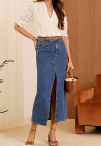 Blusa bianca in pizzo leggermente strutturata con maniche corte, abbinata a una gonna in denim a vita alta, di lunghezza al ginocchio, con uno spacco laterale e dettaglio dei bottoni.