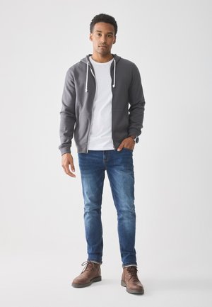 Jonge man die met één hand in zijn zak staat, gekleed in een grijze zip-up hoodie, wit T-shirt, blauwe spijkerbroek, bruine laarzen en een horloge, op een witte achtergrond.