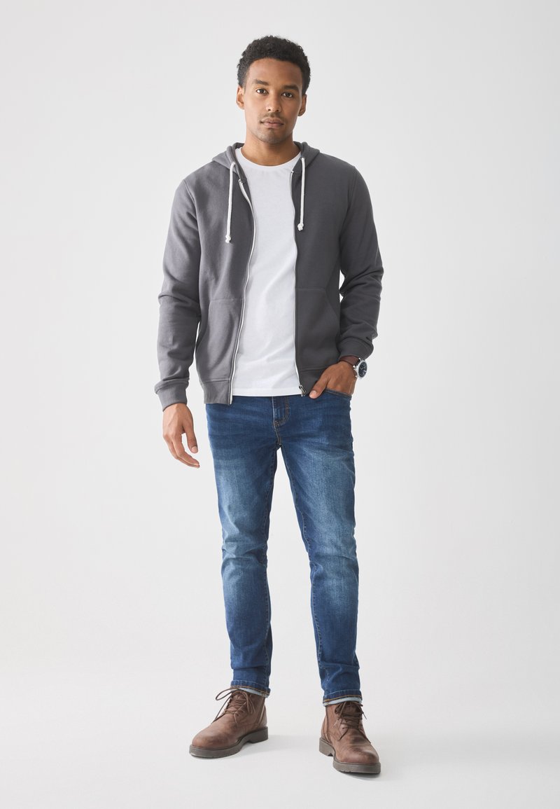 Young man standing with one hand in pocket, wearing gray zip-up hoodie, white t-shirt, blue jeans, brown boots, and wristwatch on white background.