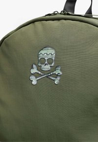 Mochila verde con tejido texturizado, que presenta un destacado diseño de calavera y tibias en un material reflectante con patrón.
