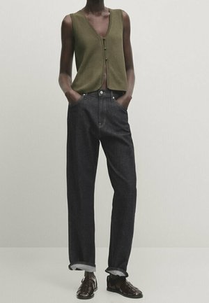 Person wearing an olive green sleeveless buttoned vest, black high-waisted straight jeans with cuffs, and black woven leather sandals.