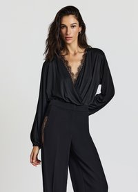 Schwarze Bluse mit tiefem V-Ausschnitt und Spitzenbesatz, langen Ärmeln und weiter Passform. Kombiniert mit hochgeschnittenen schwarzen Hosen mit Spitzenakzenten.