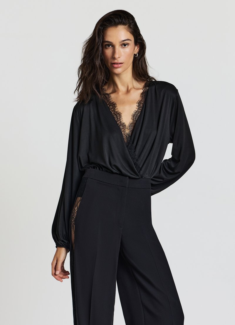 Schwarze Bluse mit tiefem V-Ausschnitt und Spitzenbesatz, langen Ärmeln und weiter Passform. Kombiniert mit hochgeschnittenen schwarzen Hosen mit Spitzenakzenten.
