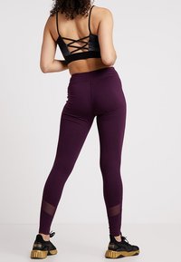 Reggiseno sportivo nero con dettagli incrociati sulla schiena, abbinato a leggings viola con pannelli in rete, indossati con scarpe da ginnastica nere e dorate.