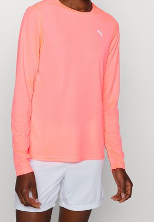 Person trägt ein langärmliges neonoranges Puma-Shirt und weiße Sportshorts vor einfachem Hintergrund.