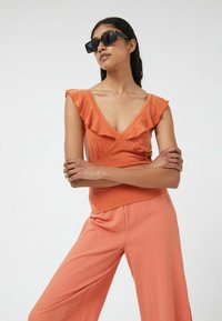 Top sin mangas naranja con un escote con volantes y un profundo corte en V, combinado con pantalones naranjas de corte holgado, ambos con una textura suave.
