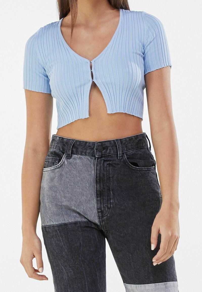 Top corto a righe azzurro chiaro con maniche corte e scollatura a V, caratterizzato da una chiusura frontale con bottoni. Indossato con jeans patchwork neri e grigi.