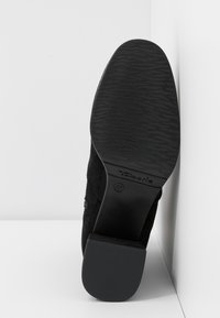 Semelle de chaussure noire avec grip texturé, taille 37, et un petit talon bloc posé à la verticale contre un fond blanc d'angle.