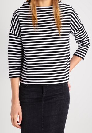 Frau trägt ein schwarz-weiß horizontal gestreiftes Langarmshirt und einen hoch taillierten schwarzen Denimrock, eine Hand in der Tasche.