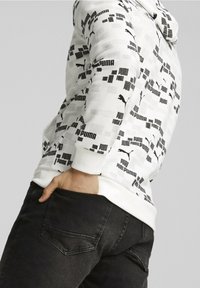 Felpa bianca con grafiche nere dei loghi Puma e motivi geometrici, abbinata a jeans di denim neri. Le caratteristiche principali includono una tasca a marsupio.