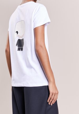 Osoba nosící bílé tričko s malou kreslenou postavičkou s bílými vlasy a černým oblečením vytištěnou na zádech, doplněné tmavými kalhotami.