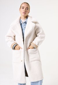 Manteau blanc en peluche avec un col à cran, fermeture à boutons à l'avant et deux grandes poches plaquées. Doublé en denim et doté de poignets marron.