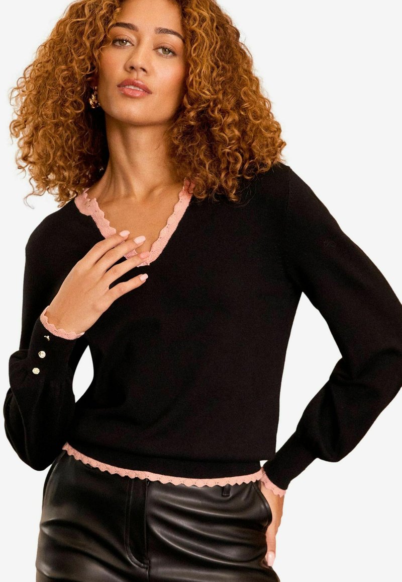 Svart tröja med V-hals, scallopformad rosa kant vid kragen och ärmsluten, samt dekorativa knappar på ärmen. Strukturerad yta.