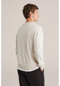 Pull en coton beige clair avec un col rond, des poignets et un ourlet côtelés, vu de dos, assorti à un pantalon foncé.